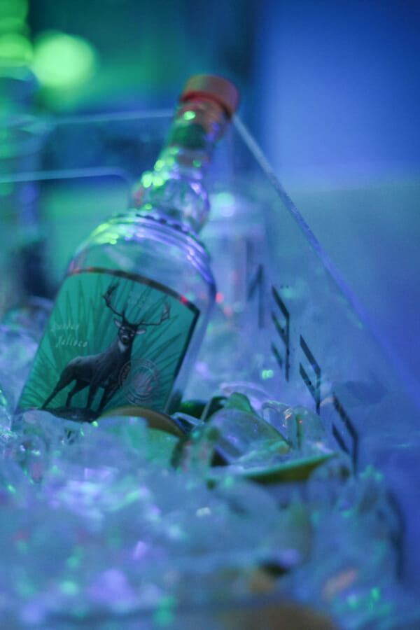 A clear tequila bottle bearing a deer-crest label rests in a bin of ice bathed in blue and green nightclub light.