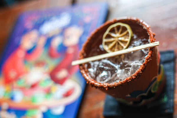 Top-down view of a red chili-salt-rimmed cocktail in a painted clay cup with a dried lime slice on top and ice inside.