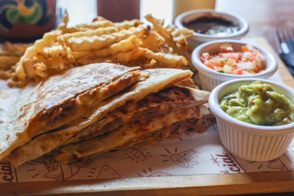 Stacked grilled quesadilla wedges beside seasoned waffle fries and three ramekins of guacamole, pico de gallo, and dark salsa.