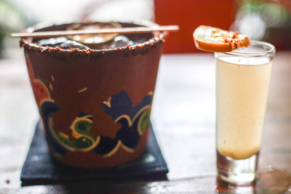 A chili-salt-rimmed drink in a painted terracotta cup beside a tall shot glass topped with a dried citrus wheel, backlit.