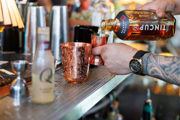 A tattooed hand tips a Tincup bourbon bottle into a copper jigger beside an ornate copper cup on a bar mat.