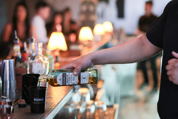 An arm pours a Bushmills Prohibition Recipe bottle into a black Bushmills cup along a lamp-lit bar with patrons behind.