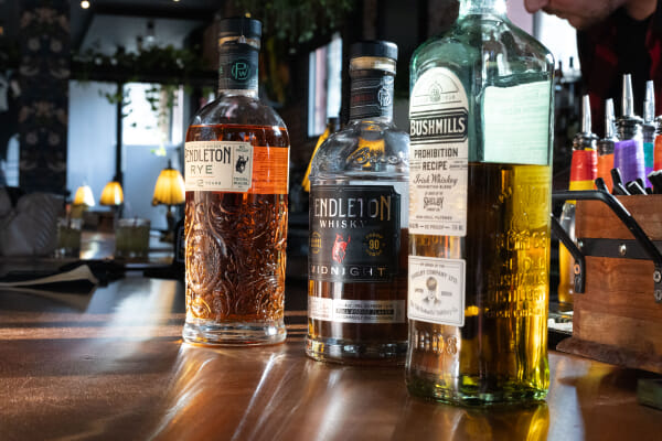 Three whiskey bottles, Pendleton Rye, Pendleton Midnight, and Bushmills Prohibition Recipe, standing in a row on a polished wood bar.