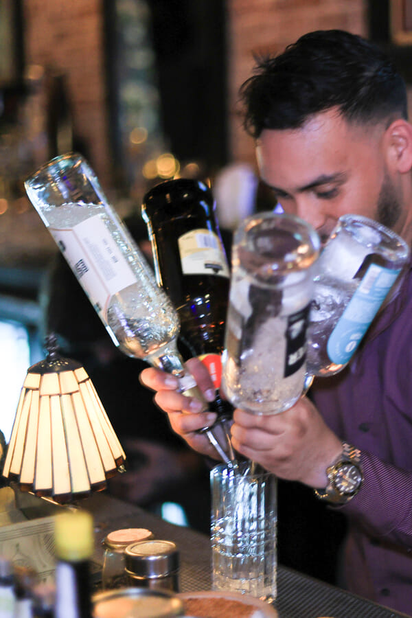 A bartender tips two upside-down liquor bottles to pour simultaneously into a tall glass behind a dimly lit bar.