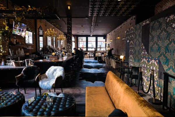 Interior of a dark upscale bar with exposed brick walls, floral wallpaper, crystal chandeliers, a long wood-topped bar with stools, and a velvet couch.