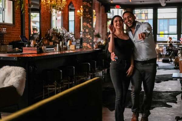 A woman in a black top and a man in a white shirt smile with arms around each other beside a dark wood bar with stools.