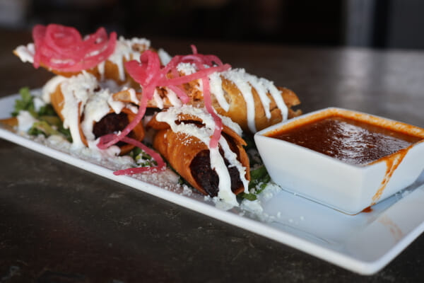 A white plate of crispy fried taquitos drizzled with crema and crumbled cotija cheese, topped with pickled red onions, beside a square dish of red salsa.